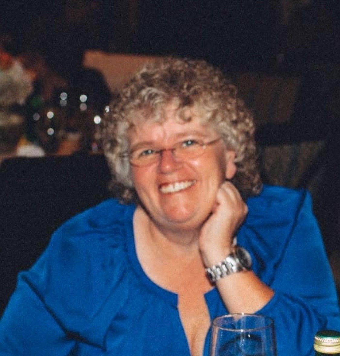 Smiling woman with short curly gray hair and glasses, wearing a blue blouse, resting her chin on her hand at a table with a wine glass nearby.