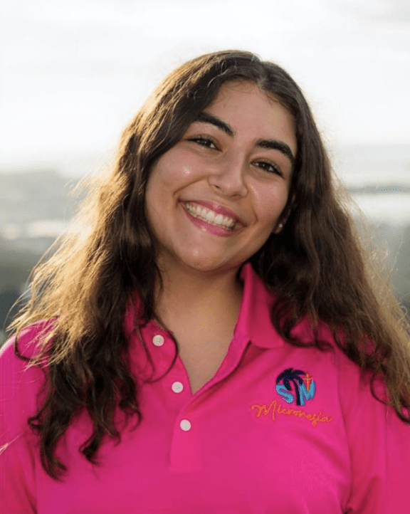 Young woman with long dark hair smiling at the camera, wearing a bright pink polo shirt with a logo on the left chest.