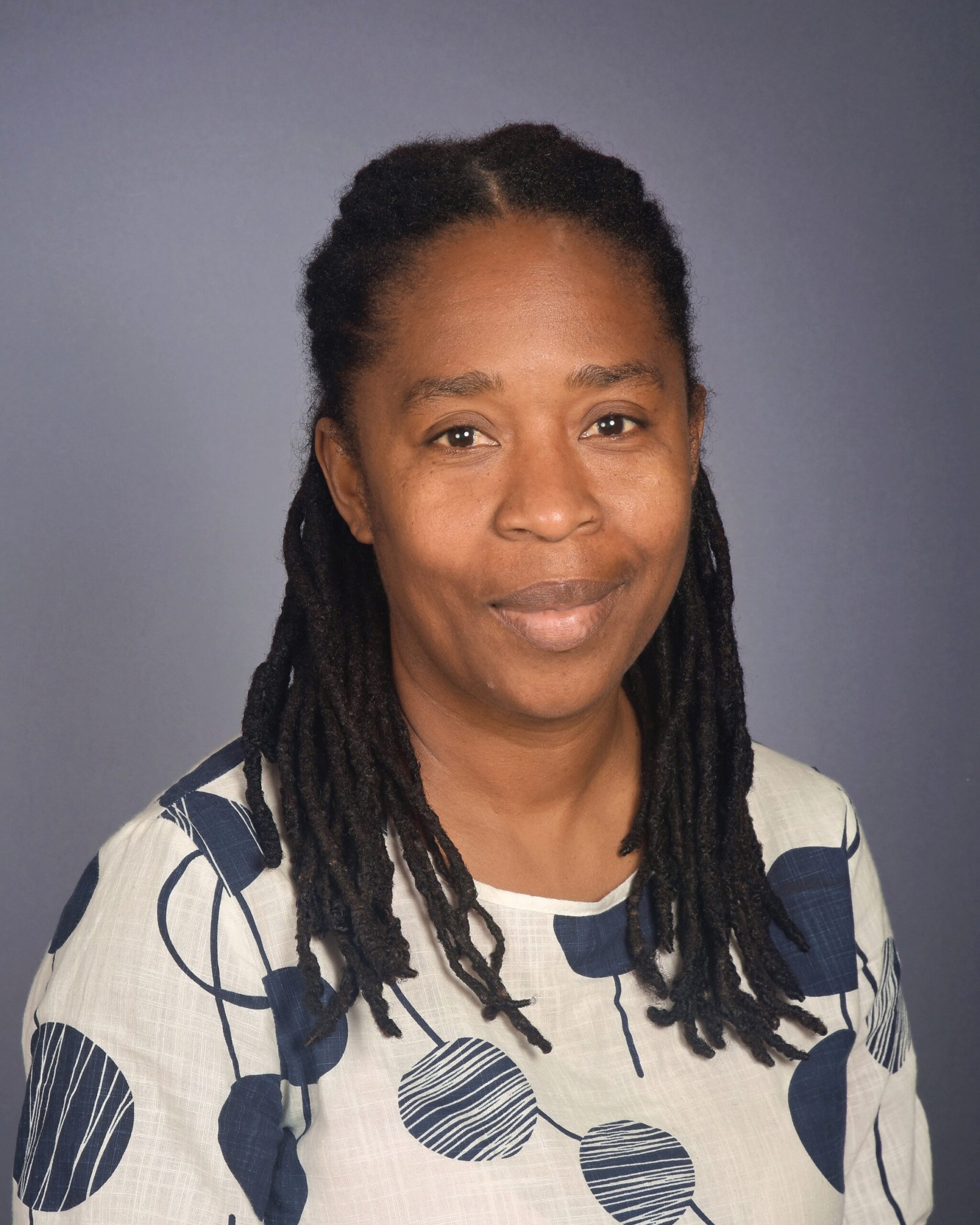 Portrait of a woman with long dreadlocks, wearing a light patterned blouse, smiling softly against a gray backdrop